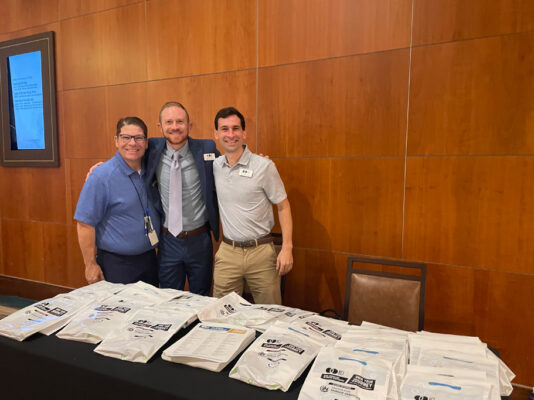 Three men standing behind table of conference bags